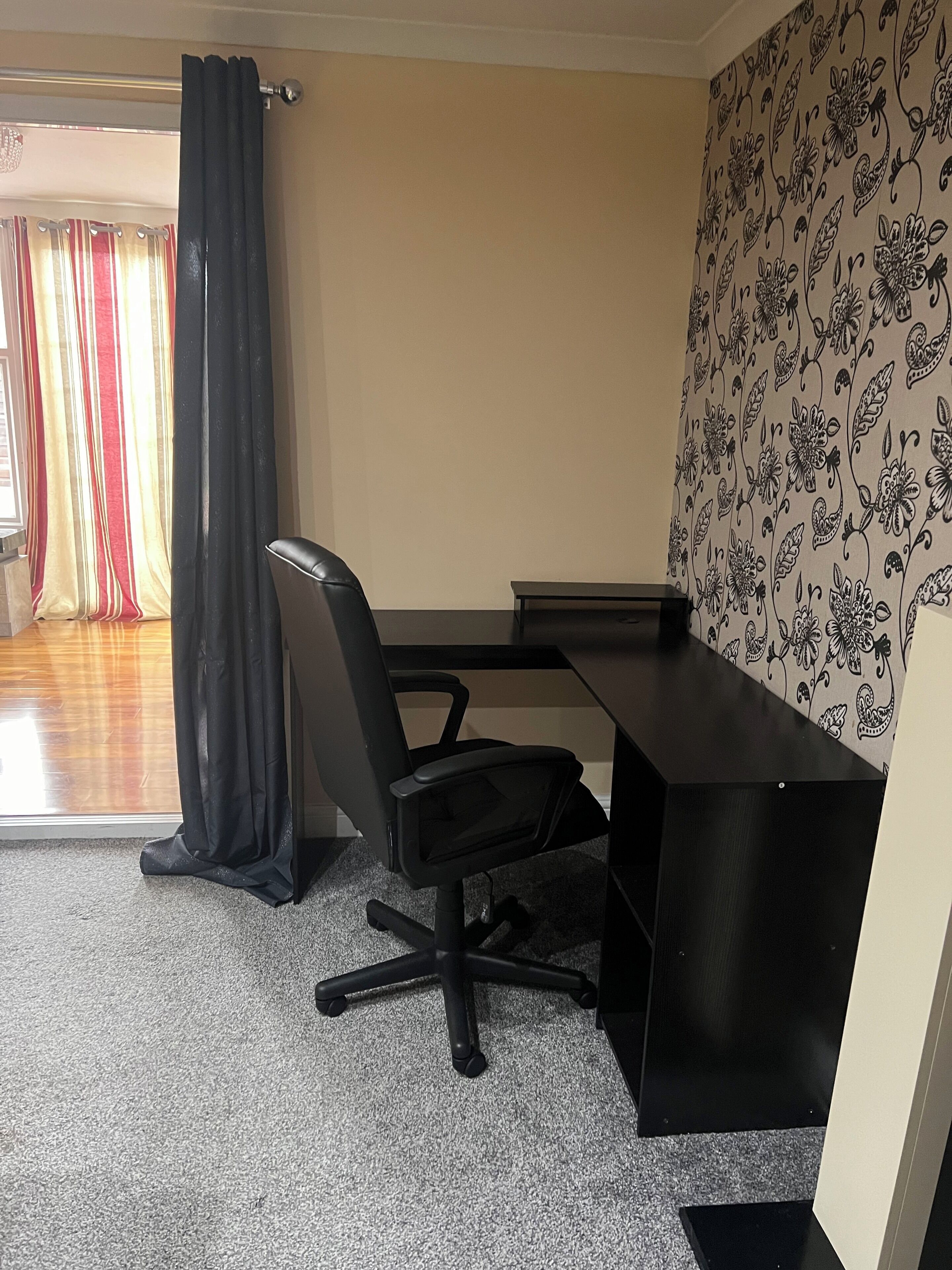 A room with a desk, a black office chair, a patterned wallpaper, and a window with curtains.