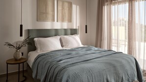 A modern bedroom with a bed, a round side table, a vase with branches, and two wall-mounted artworks.