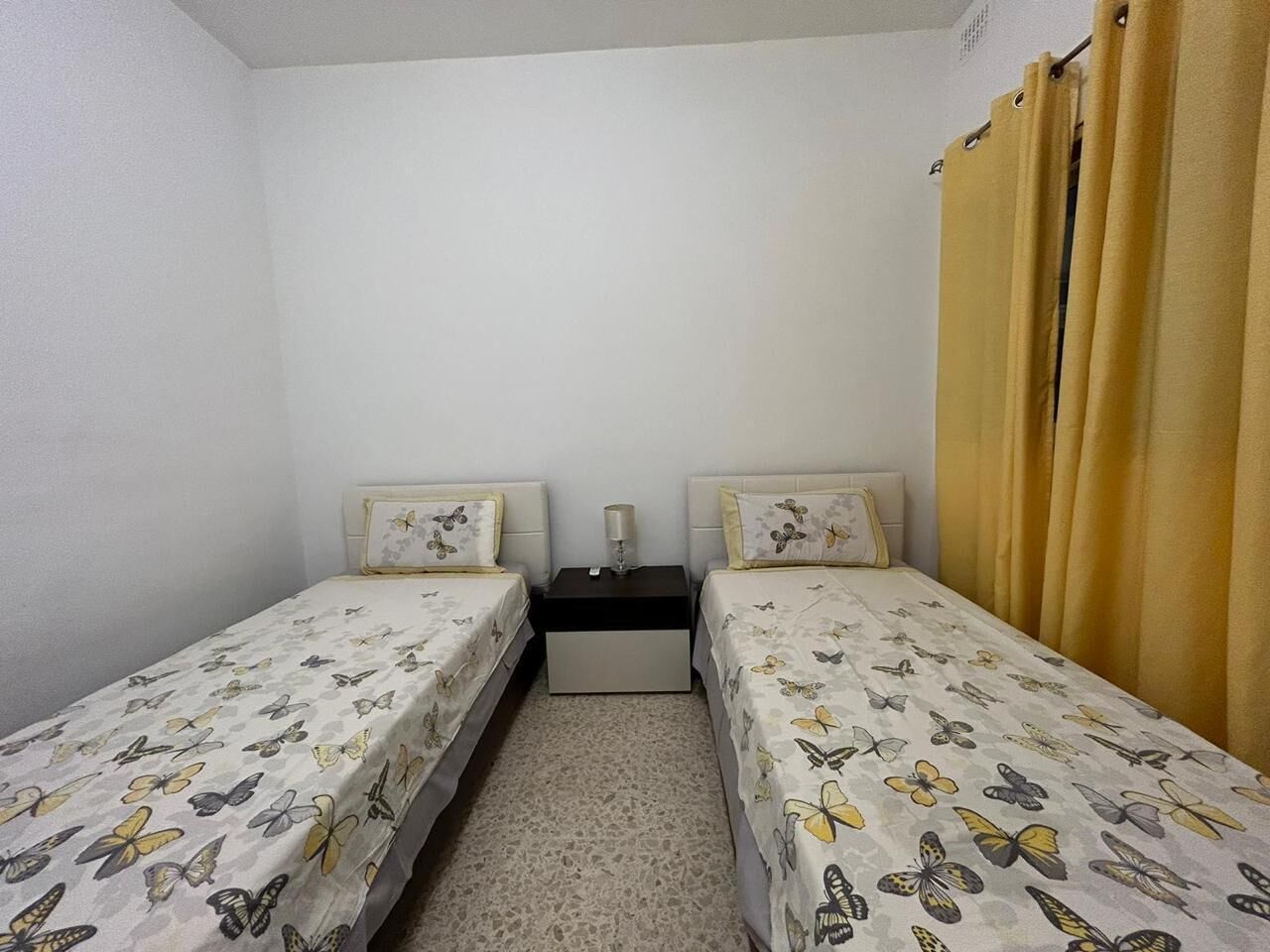 Une petite chambre individuelle avec deux lits simples, une table de chevet et une fenêtre ornée de rideaux jaunes.
