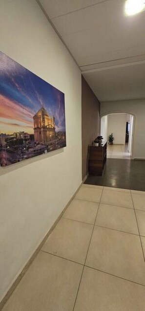 A hallway with a tiled floor, a wall-mounted painting of a building, and a small wooden table with a plant.