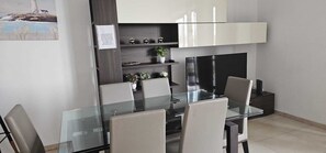 A modern dining area with a glass table and chairs, a dark wooden cabinet above, and a small shelf with decorative items.