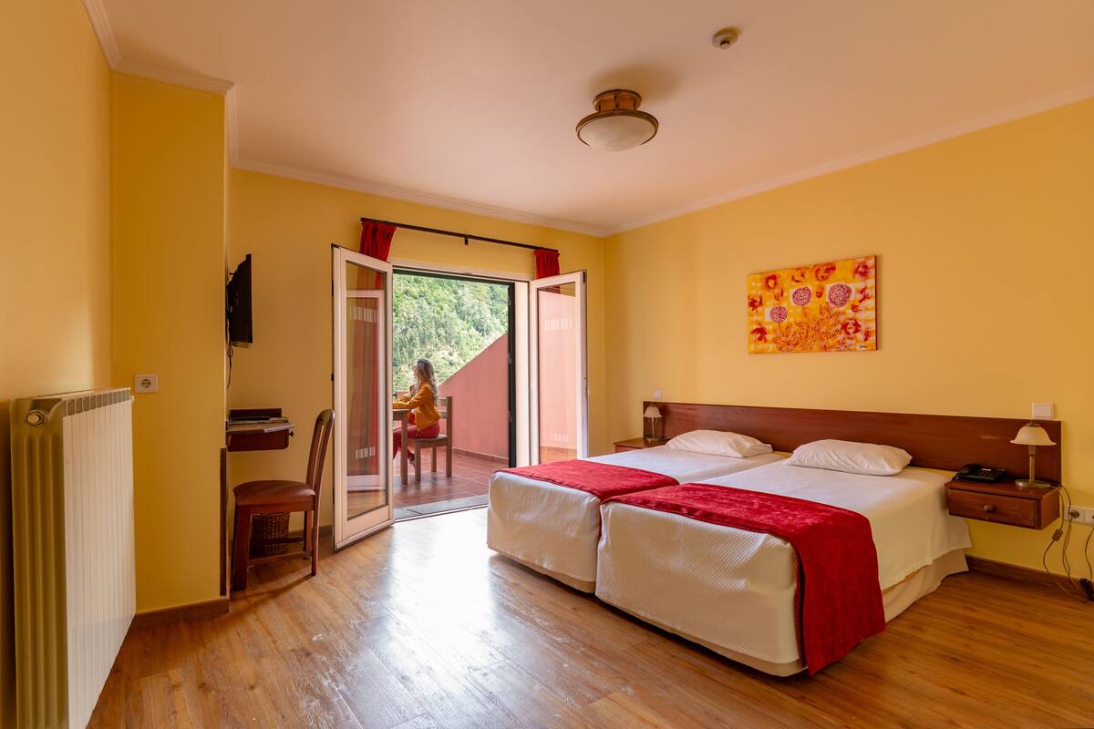 A double bed with a red blanket, a wooden nightstand, a wall-mounted TV, and a sliding glass door leading to an outdoor area.