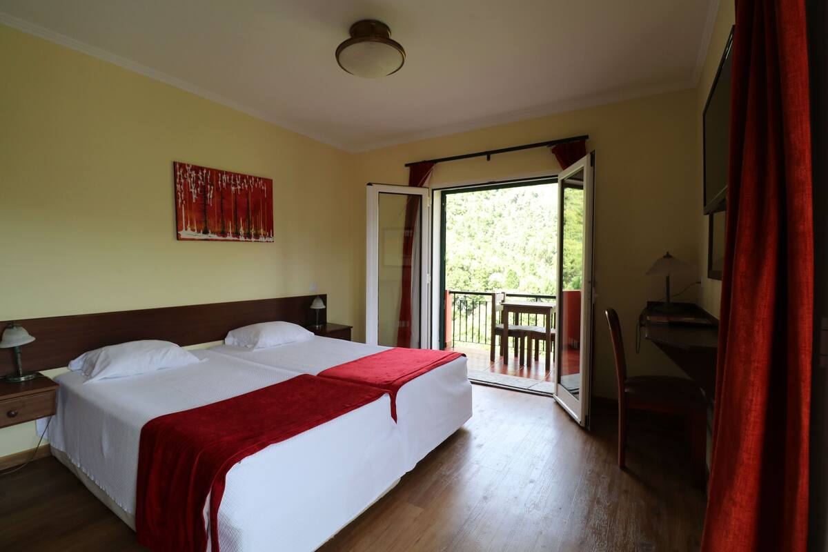 A hotel room with a large bed, a desk, a chair, and a balcony with a view of greenery.