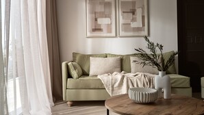 En stue med en grøn sofa, to puder, en hvid vase med grene og et rundt træbord.