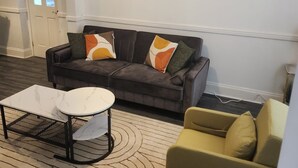 Ein modernes Wohnzimmer mit einem grauen Sofa, einem kleinen weißen Couchtisch und einem gelben Sessel. An der Wand hängen zwei gerahmte Bilder, eines zeigt einen Elefanten, das andere eine Landschaft.