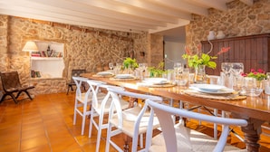 A dining area with a long wooden table set for six, surrounded by white chairs. A stone wall and a wooden cabinet with a shelf are visible in the background. - L'ERESTA - Wonderful villa with private pool and Free Wifi (Pollença)