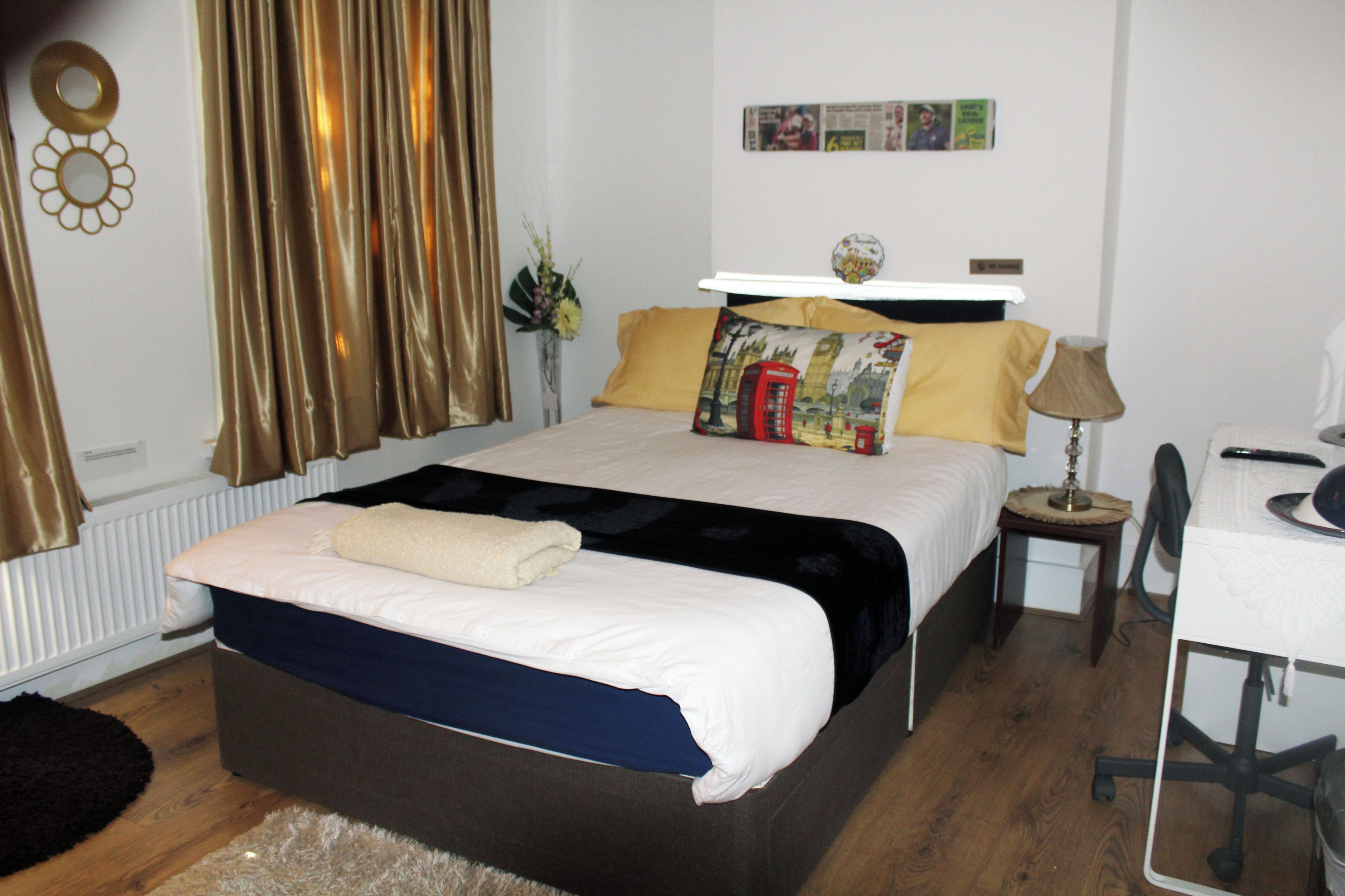 A neatly made bed with a white duvet, a black throw, and a yellow pillow. There is a small bedside table with a lamp, a chair, and a wall-mounted shelf above the bed.
