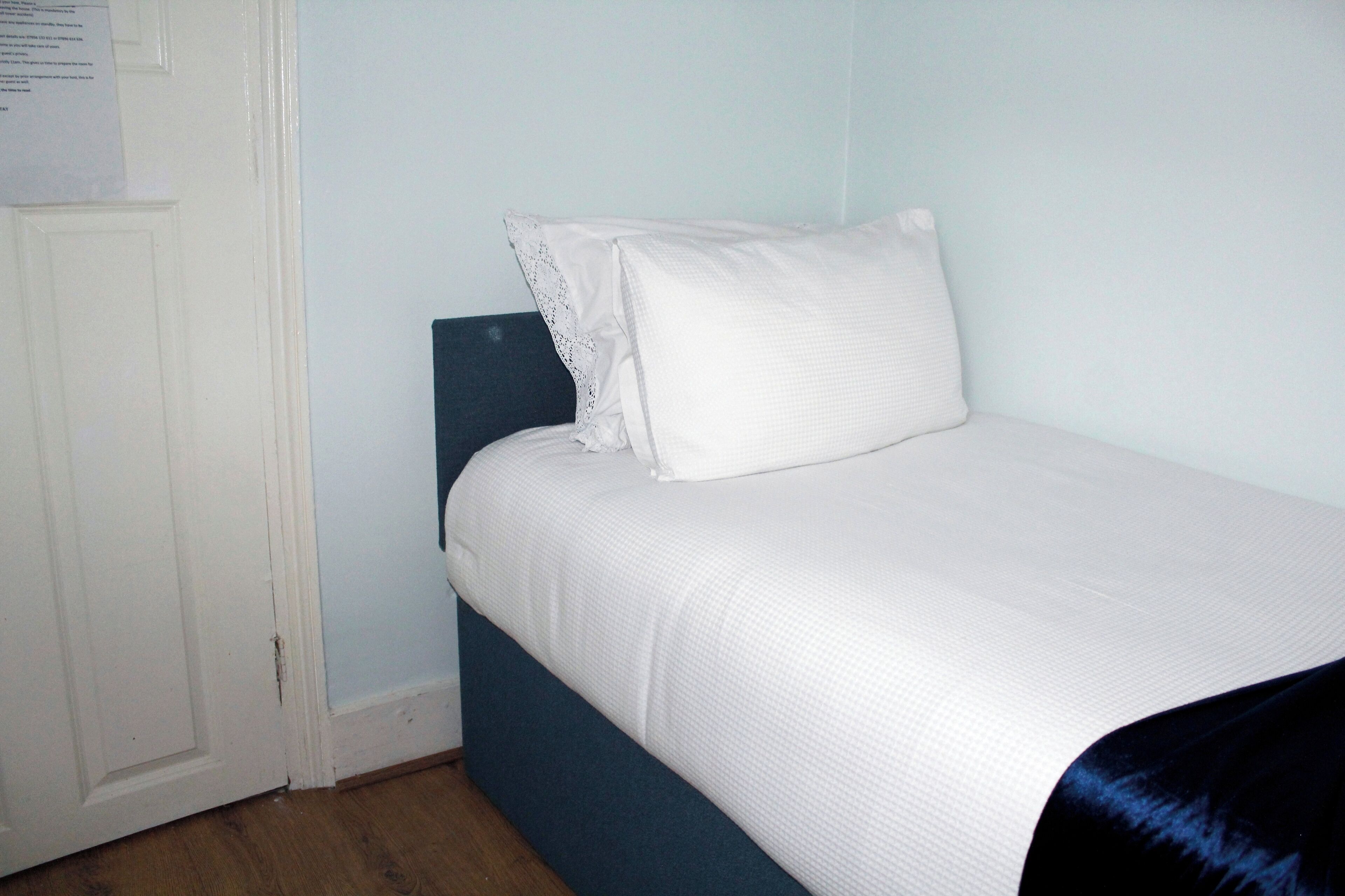 Un lit simple avec un matelas et un oreiller blancs, une tête de lit bleu foncé et une porte blanche sur laquelle est collé un petit morceau de papier.