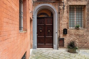 A traditional brick building with a large wooden door, a small window with a grid pattern, and a potted plant on the ground. - Palazzo Patrizi Suite & Spa  (Siena)