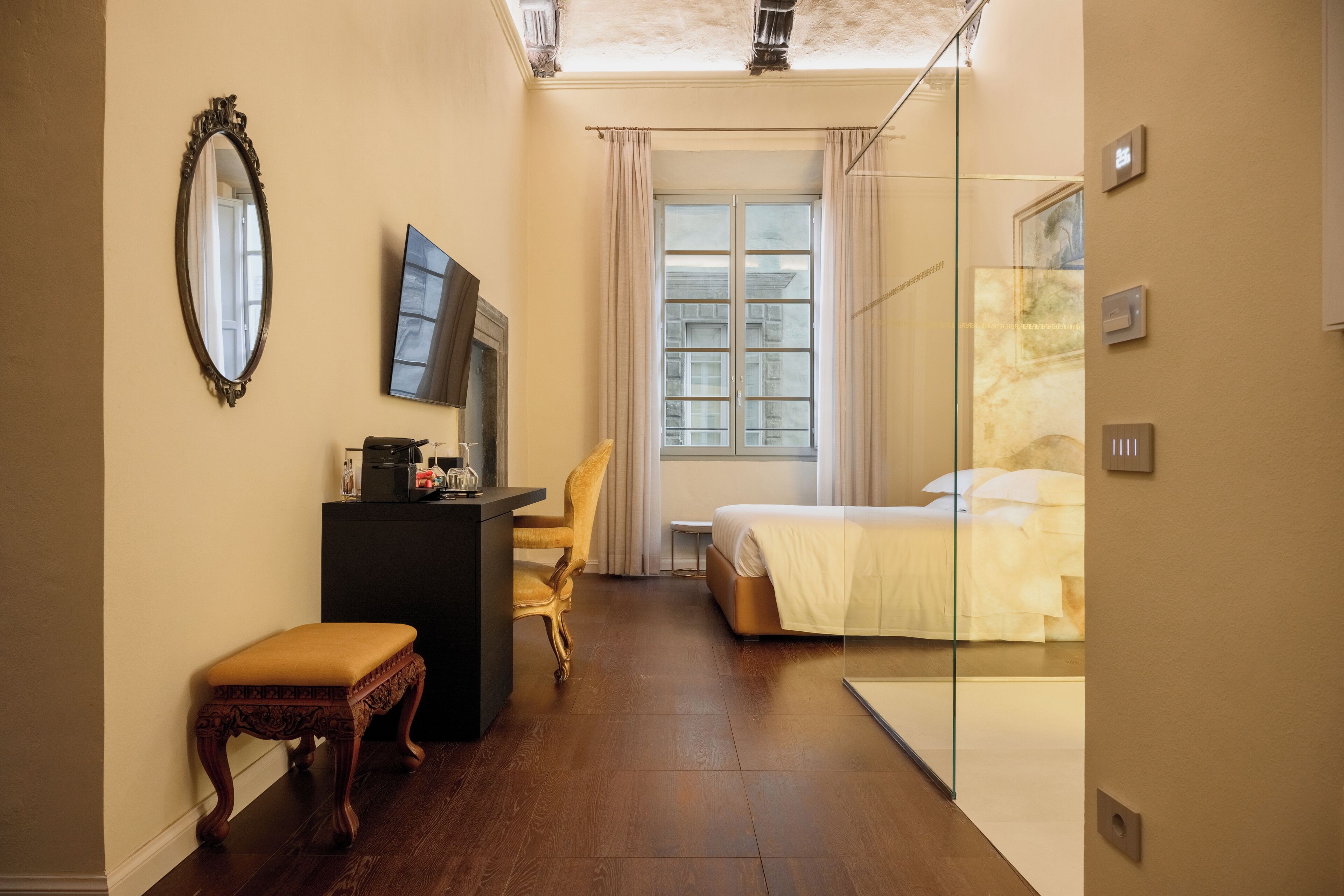 Een kleine hotelkamer met een bed, een bureau met stoel, een tafeltje, een spiegel en een raam met gordijnen.