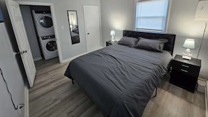 Une chambre à coucher comprenant un lit, une table de chevet, une lampe, une fenêtre, une porte et un coin buanderie.