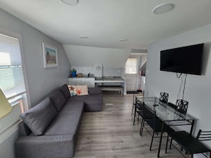 Ein modernes Wohnzimmer mit einem grauen Ecksofa, einem Esstisch mit Stühlen, einer Küche im Hintergrund und einem flachbildfernseher an der Wand.