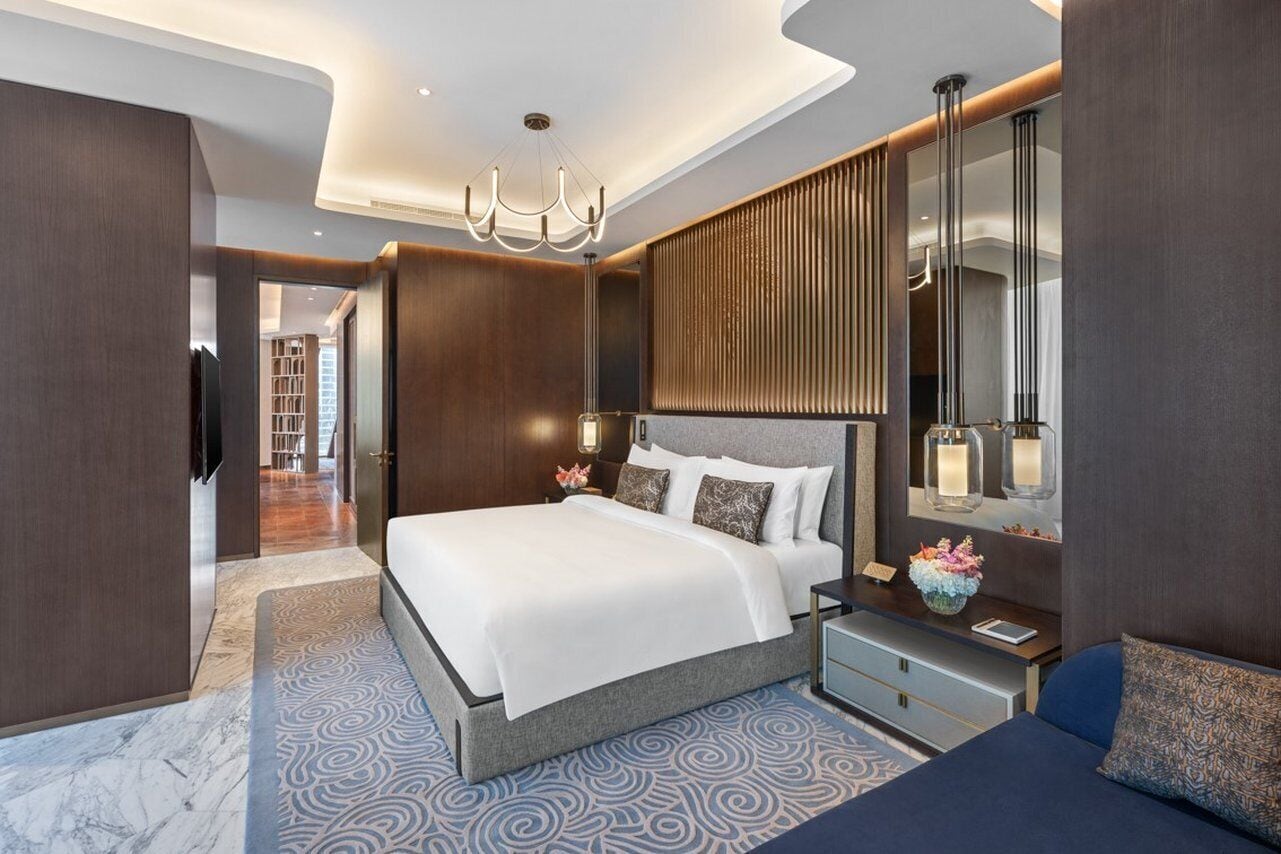 Quarto de hotel moderno com uma cama grande, uma mesinha de cabeceira, um espelho e um lustre.