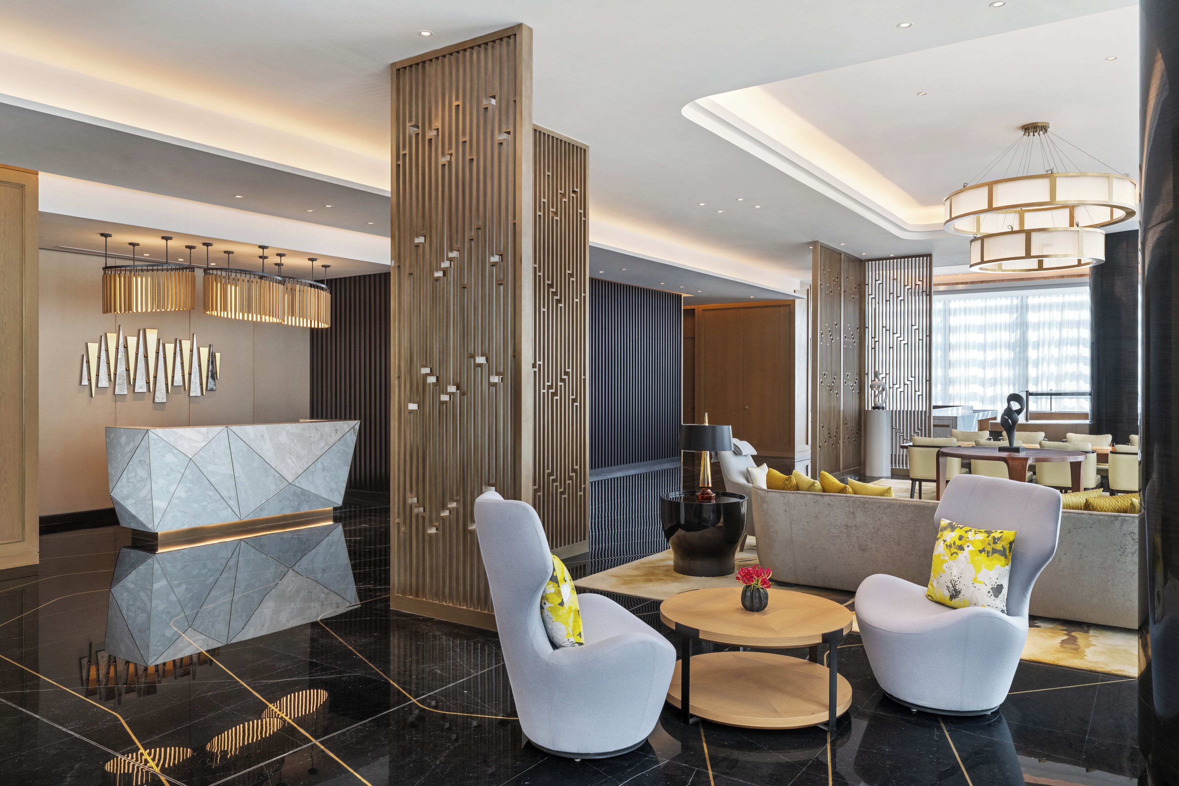 A modern lobby with a reception desk, two armchairs, a round table, and a decorative wall panel.