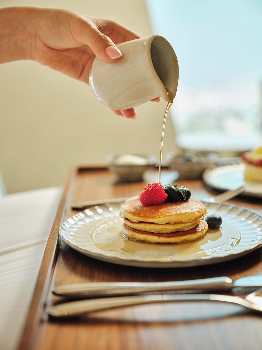 Une main verse du sirop sur des crêpes garnies de fruits rouges, le tout présenté sur une assiette.