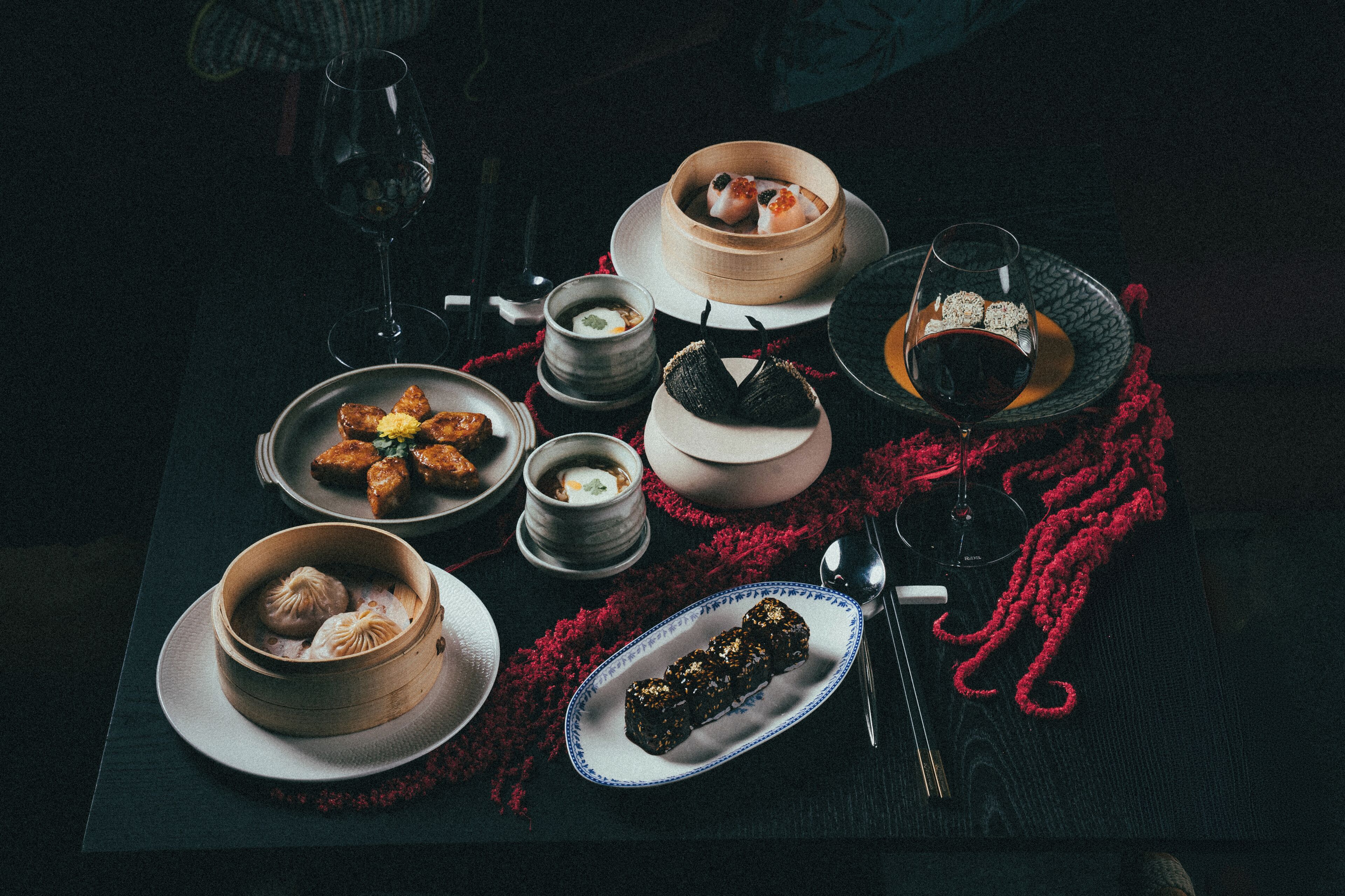 Una mesa servida con varios platos, entre ellos dumplings y una copa de vino tinto, sobre un mantel oscuro.