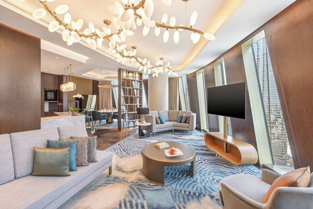 Un salon moderne avec un grand canapé, une table basse et un téléviseur. On aperçoit une bibliothèque et un coin repas au fond de la pièce.