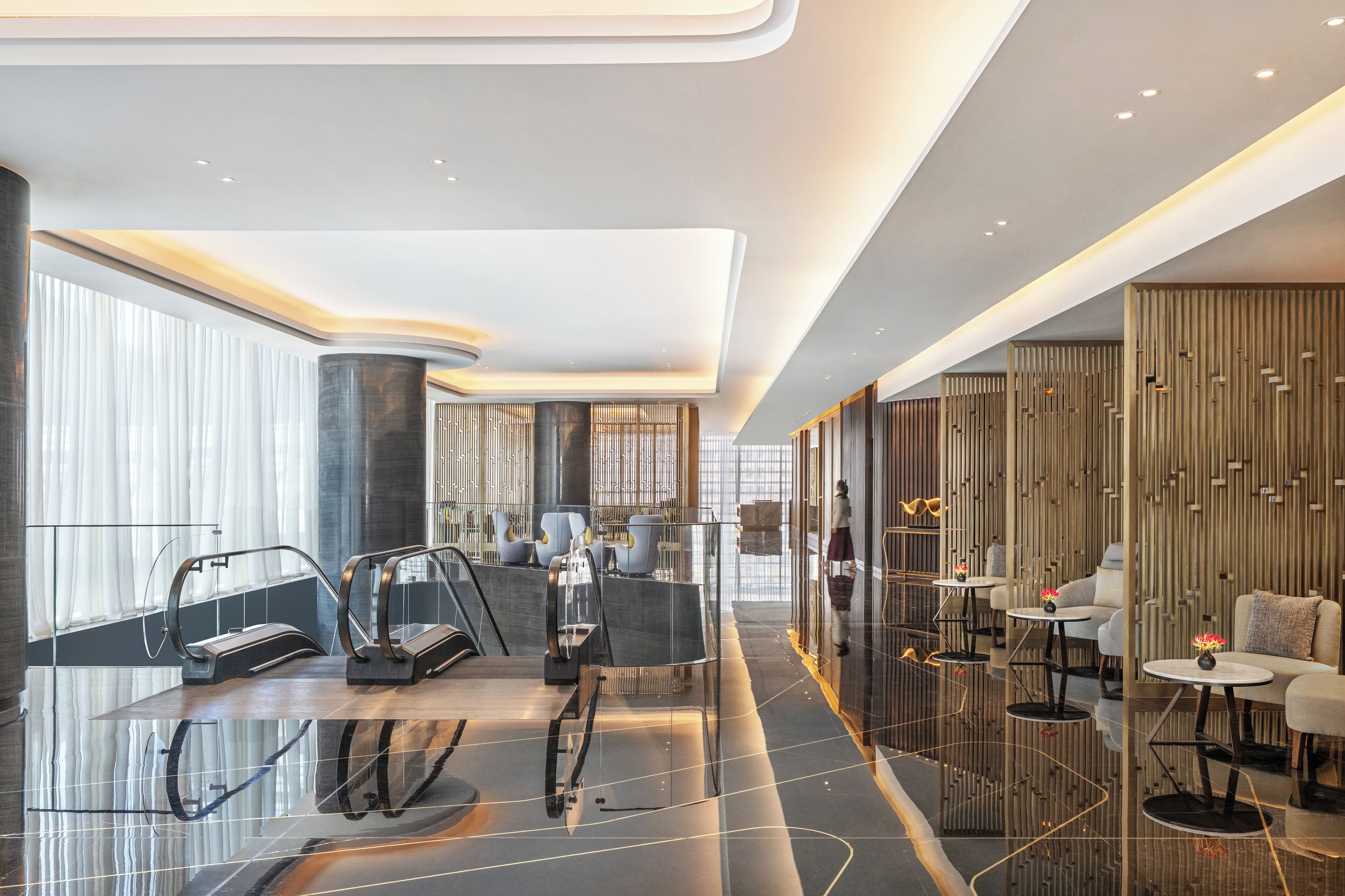 A modern hotel lobby with a glass elevator, seating areas, and a decorative wall.