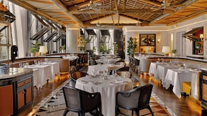 Un vaste espace dédié à la salle à manger, avec de nombreuses tables dressées, de hauts plafonds avec des poutres apparentes et de grandes fenêtres laissant entrer la lumière naturelle.