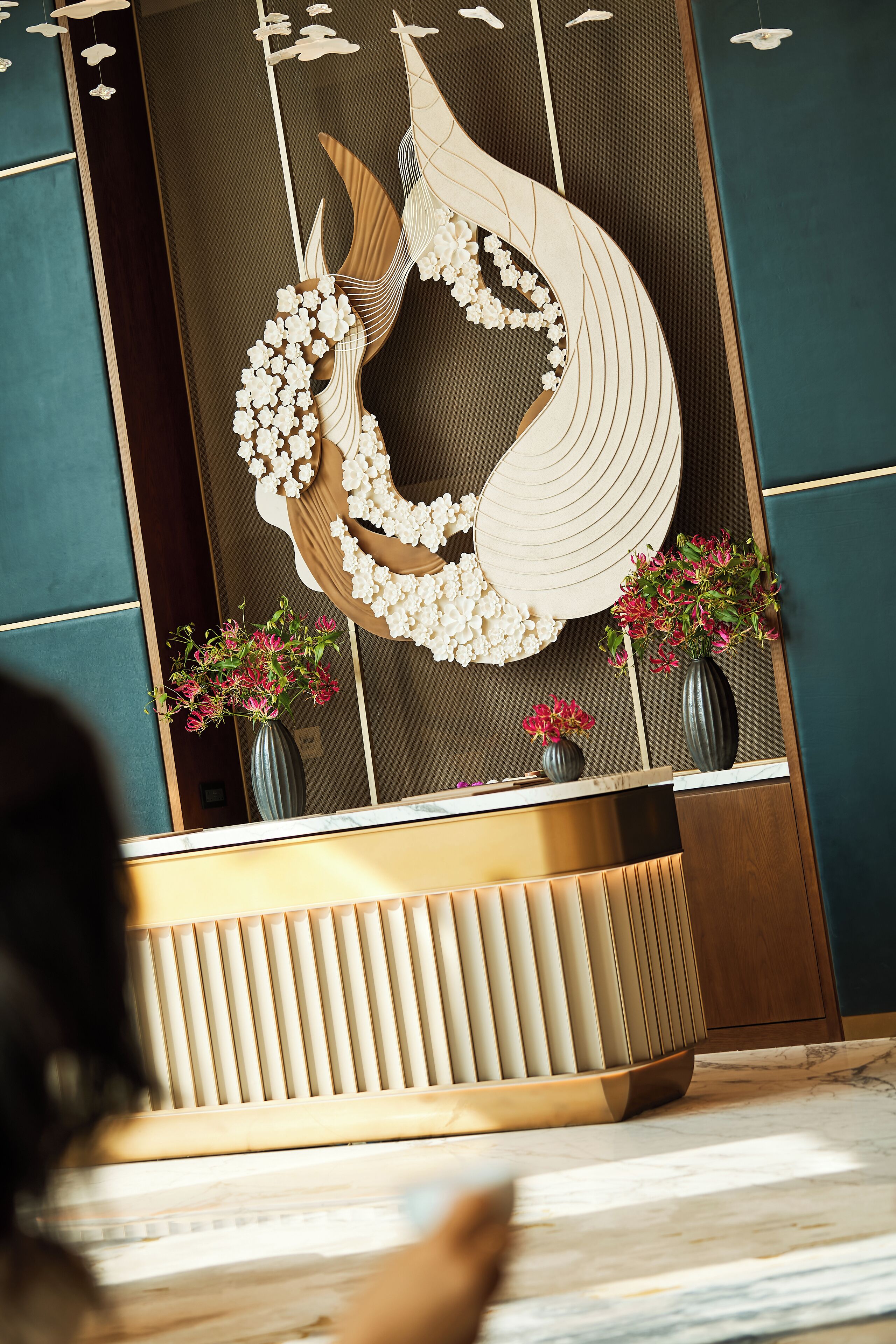 A reception desk with a modern design, a large decorative wall piece, and two vases with flowers.