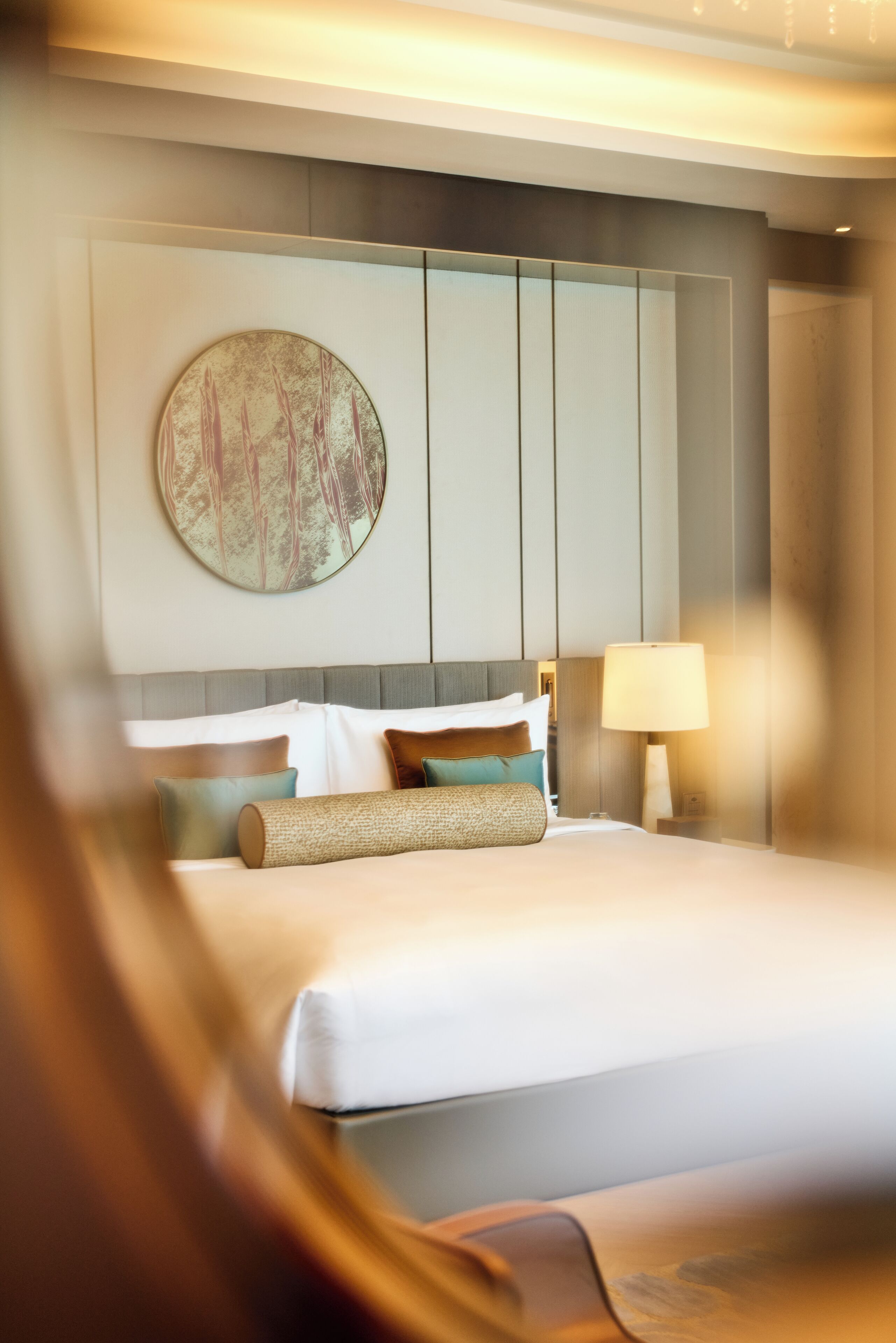 A modern hotel room with a large bed, a round wall art piece, and a nightstand with a lamp.