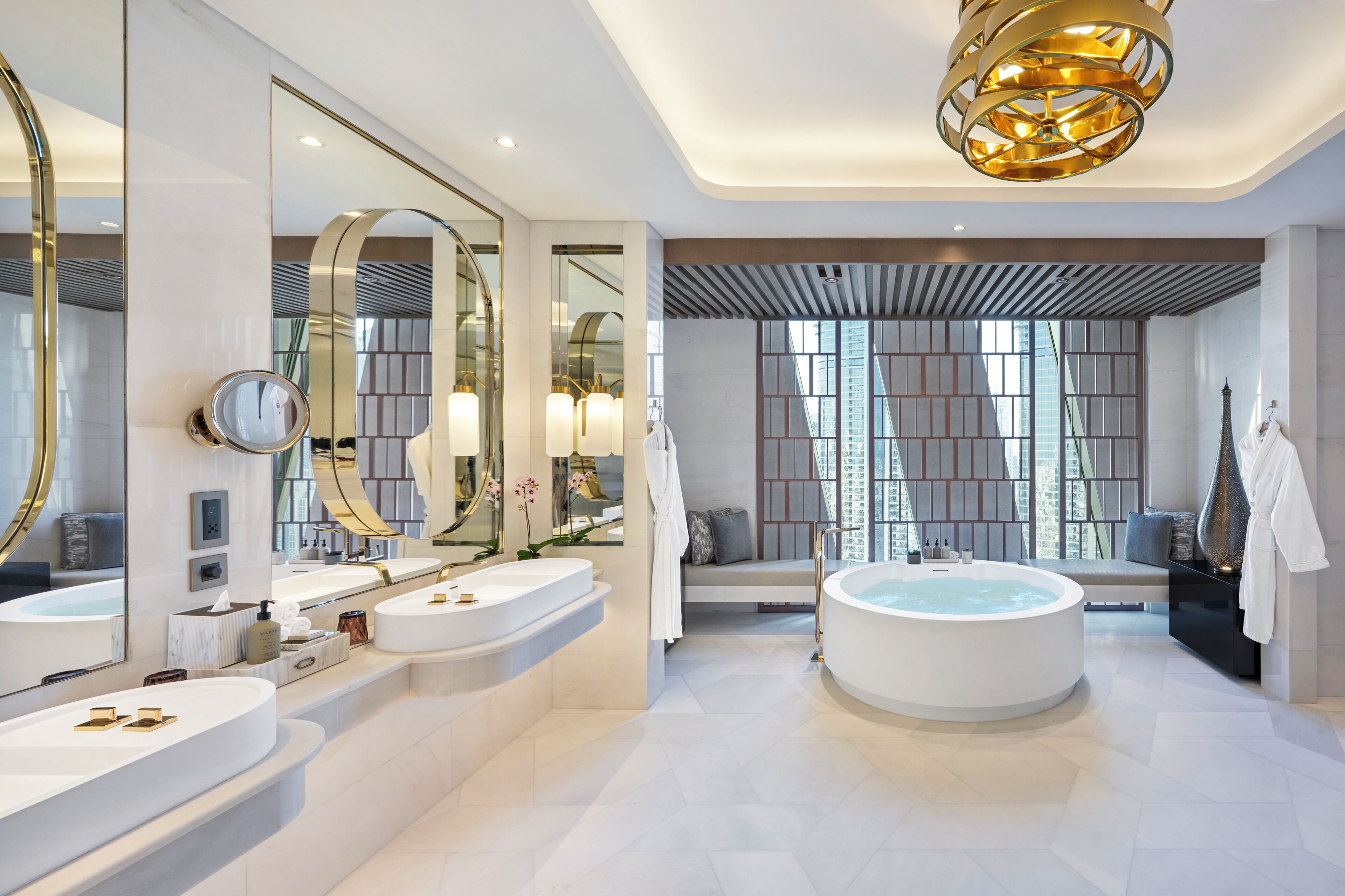 A modern bathroom with two sinks, a large bathtub, and a seating area with a view of the city.