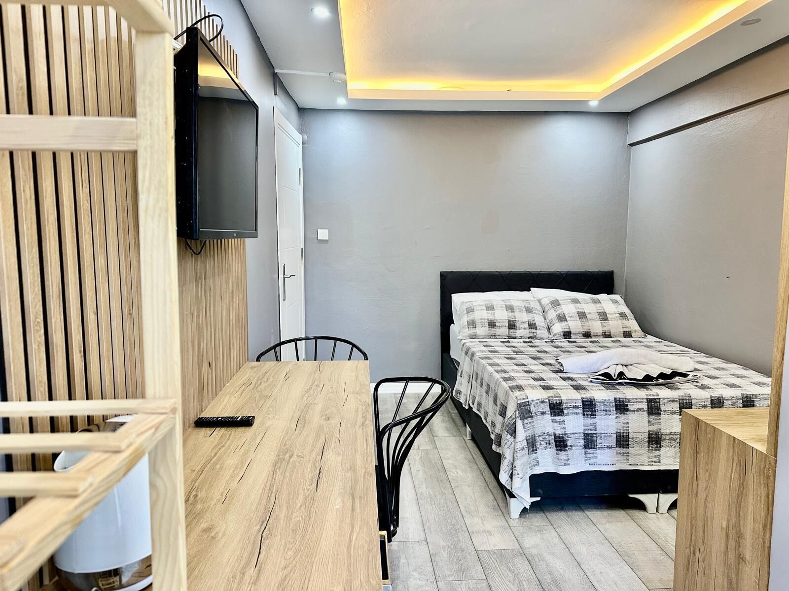 A small, modern bedroom with a bed, a wooden table, and a TV mounted on the wall.