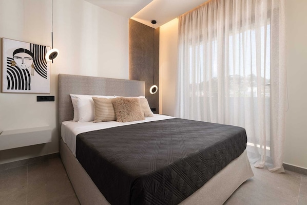Un dormitorio moderno con cama, cabecera, luz empotrada en la pared y ventana con cortinas de gasa.