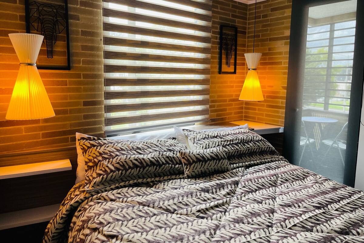 A bedroom with a bed, two wall-mounted lamps, a window with blinds, and a view of the outdoors.