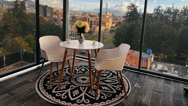 A room with a large window overlooking a cityscape, featuring a round table with chairs and a vase of flowers. - SUITES H&A 911 (Bogotá)