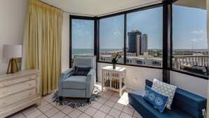 A modern room with a view of the city and ocean. It includes a blue sofa, a white dresser, and a small white side table with a vase.