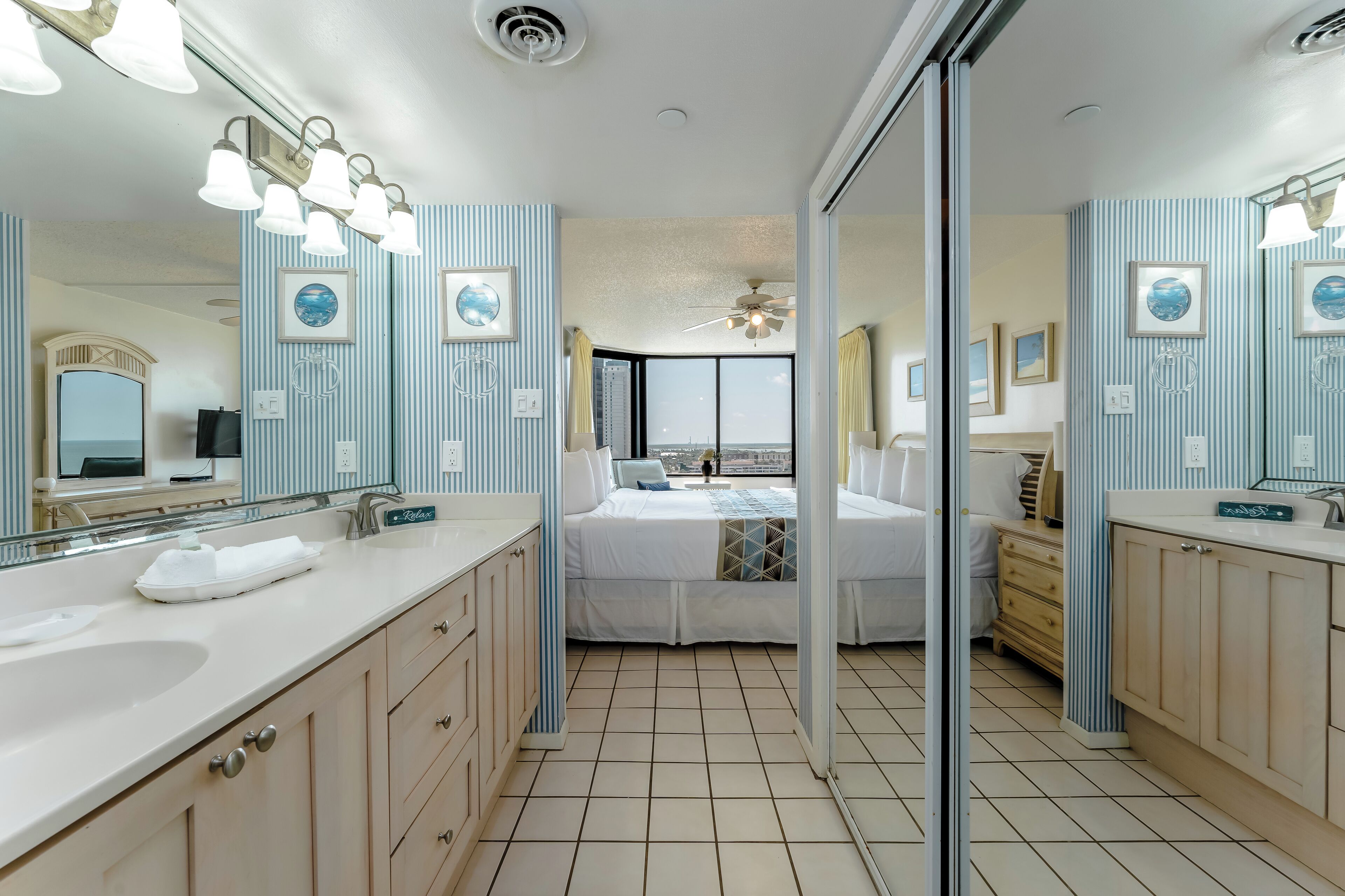 A hotel bathroom with two sinks, a large mirror, and a bed with a view of the city.