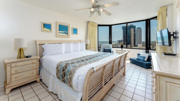 A hotel room with a large bed, a desk, a chair, a ceiling fan, and a city view.