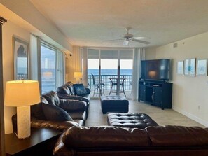 Ein Wohnzimmer mit Sofa, Hocker und Fernseher auf einem Schrank. Es gibt einen Balkon mit Tisch und Stühlen sowie Blick aufs Meer.
