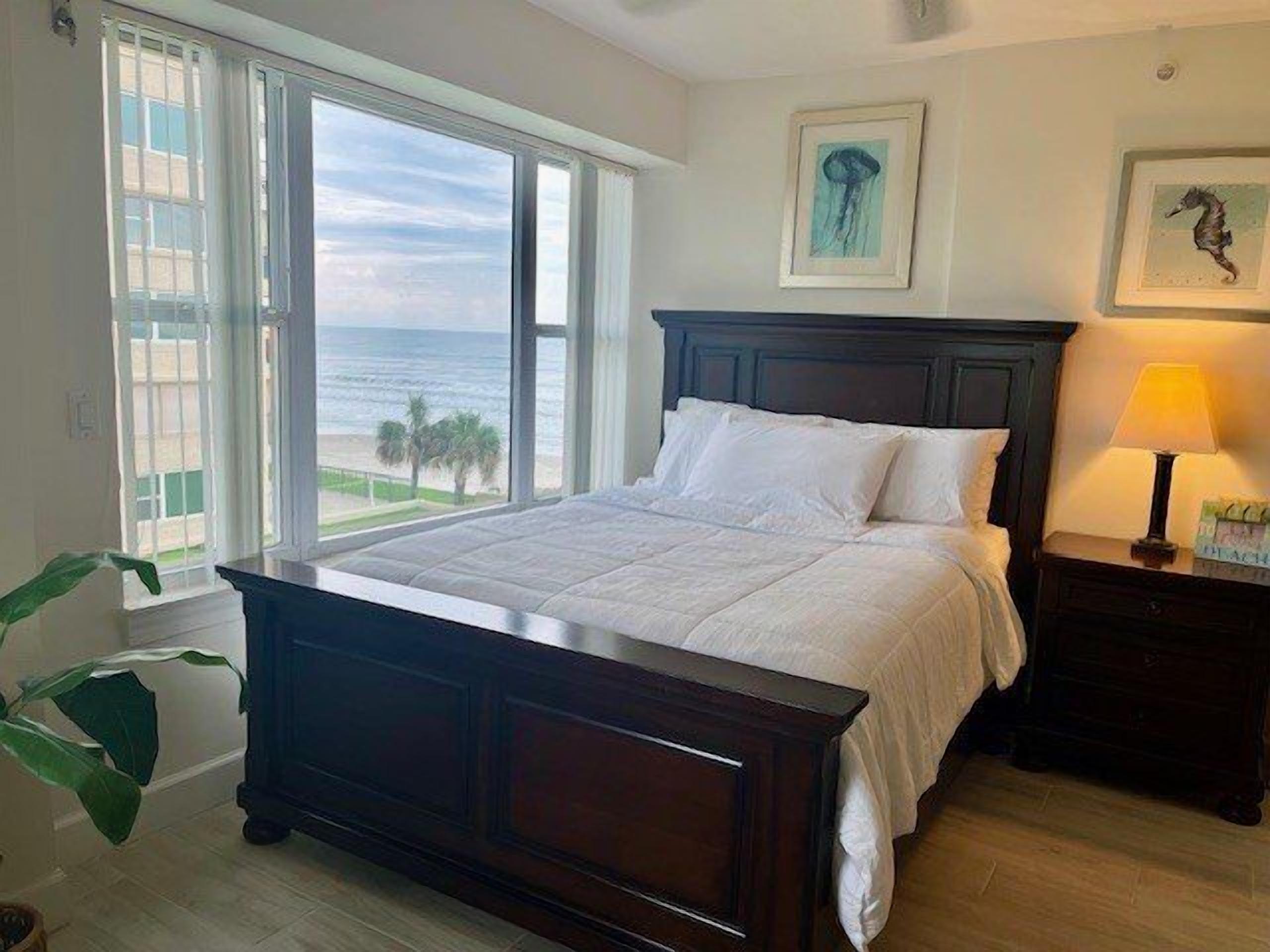 A bedroom with a large bed, a nightstand with a lamp, and a view of the ocean through large windows.