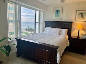 A bedroom with a large bed, a nightstand with a lamp, and a view of the ocean through large windows.