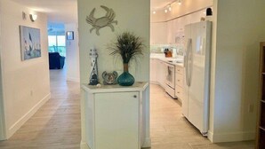 A modern kitchen with white cabinets, a built-in oven, and a refrigerator. There is a small white cabinet with decorative items on top. - Towers 8 & 9 8301 - Daytona Beach Oceanfront Vacations, Inc (Ponce Inlet)