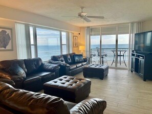 A spacious living room with a large leather sofa, a coffee table, and a television. There is a balcony with a view of the ocean. - Towers 8 & 9 8301 - Daytona Beach Oceanfront Vacations, Inc (Ponce Inlet)