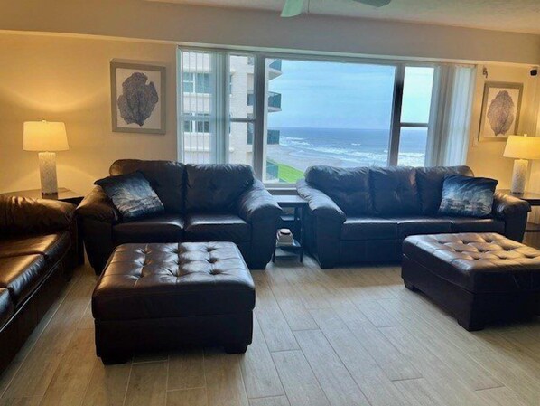 A living room with two leather sofas, a matching ottoman, and a view of the ocean through large windows. - Luxury at its Finest in this 3/3 Oceanfront #8301 at the Towers in Ponce Inlet (Ponce Inlet)