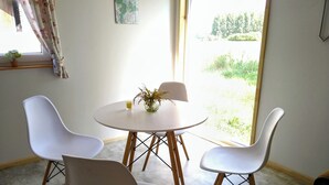 A dining area with a round table and four white chairs, a vase with flowers, and a window with a view of greenery. - Urlaub in Masuren - Ferienhaus, Angelurlaub (Iława)