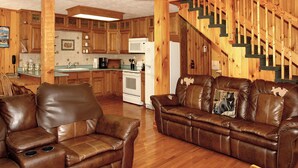 Amplia sala de estar con sofá y sillón de cuero marrón, piso de madera, área de cocina con gabinetes de madera, refrigerador blanco y escalera de madera.