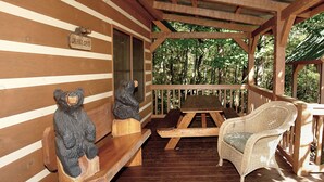 Una cabaña de madera con un porche, dos bancos de madera, una silla de mimbre y dos esculturas de osos.
