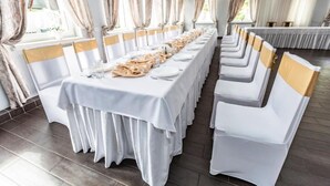 Egy hosszú, fehér terítővel beborított bankettasztal, arany színű székvédőkkel, egy nagy ablakokkal és függönyökkel ellátott teremben.