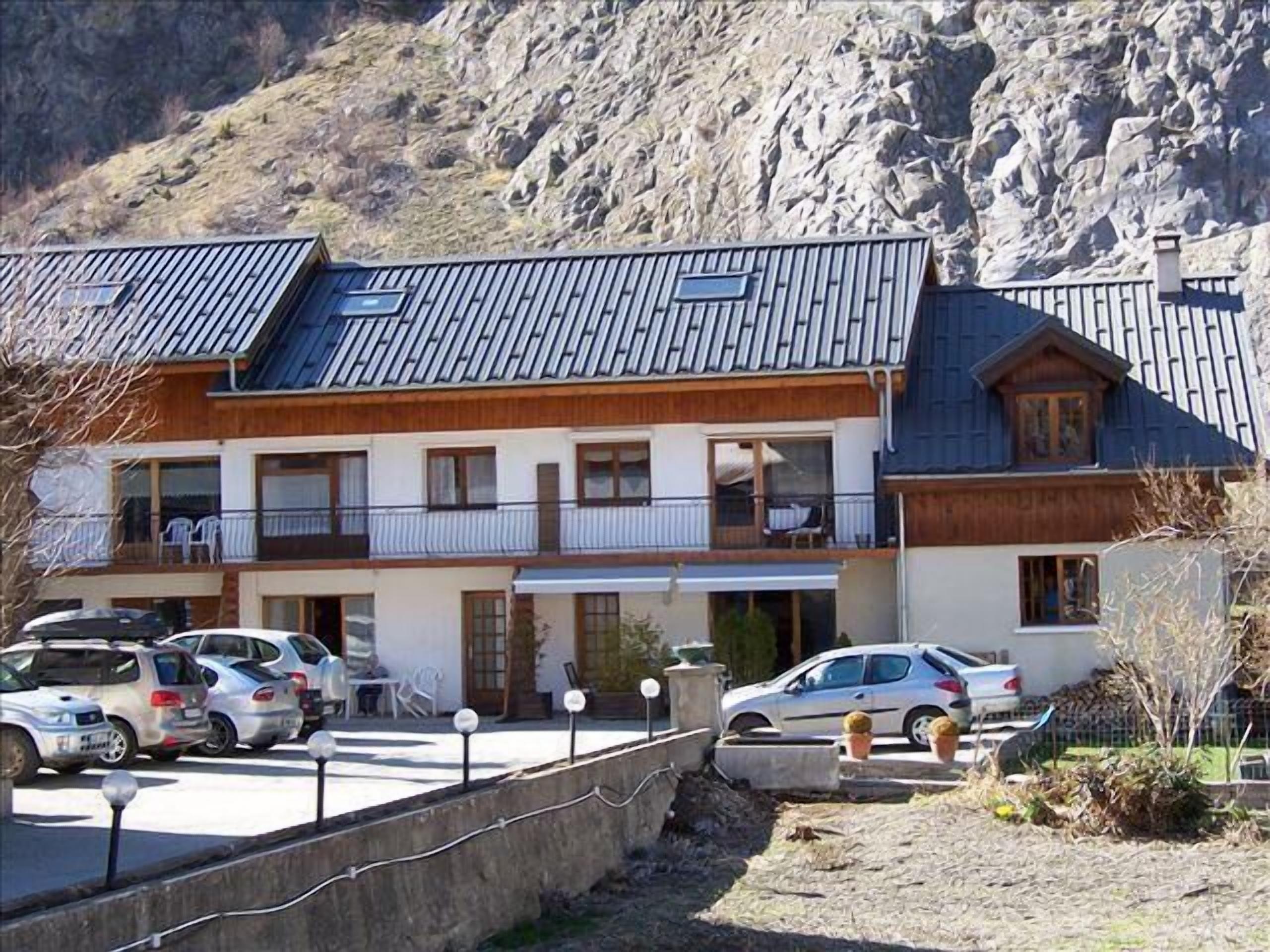 A multi-story building with a sloped roof, balconies, and a parking area with cars.