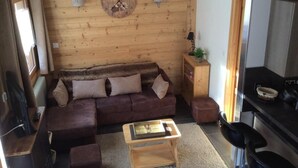 Een knus interieur van een chalet met een houten wand, een bruine bank, een houten salontafel, een keuken met een fornuis en een klok aan de muur.