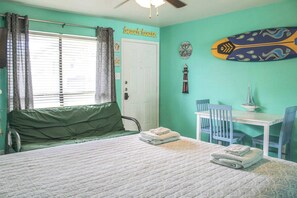 Quarto com cama, escrivaninha e cadeiras. Há uma janela com cortinas, uma porta e uma parede decorada com um quadro de surf.