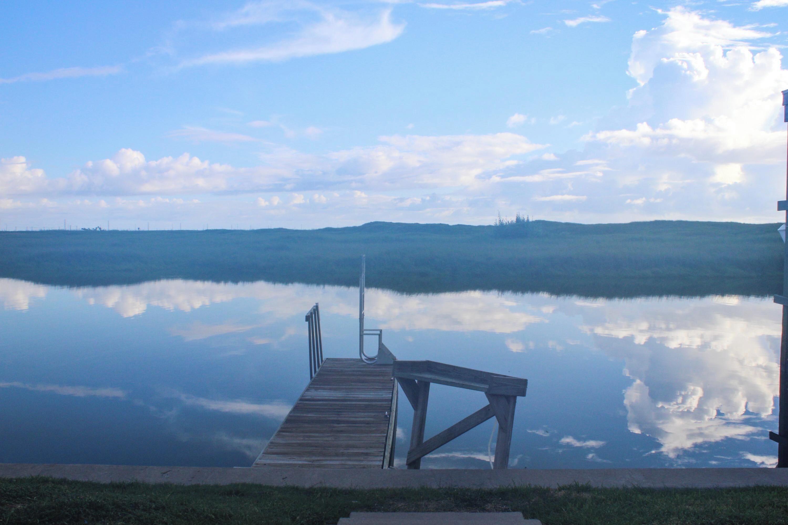 A wooden dock extending into calm water with a clear reflection of the sky and distant hills.