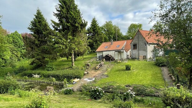 Ein rustikales Haus mit rotem Ziegeldach, umgeben von üppiger Vegetation und einem kleinen Teich.