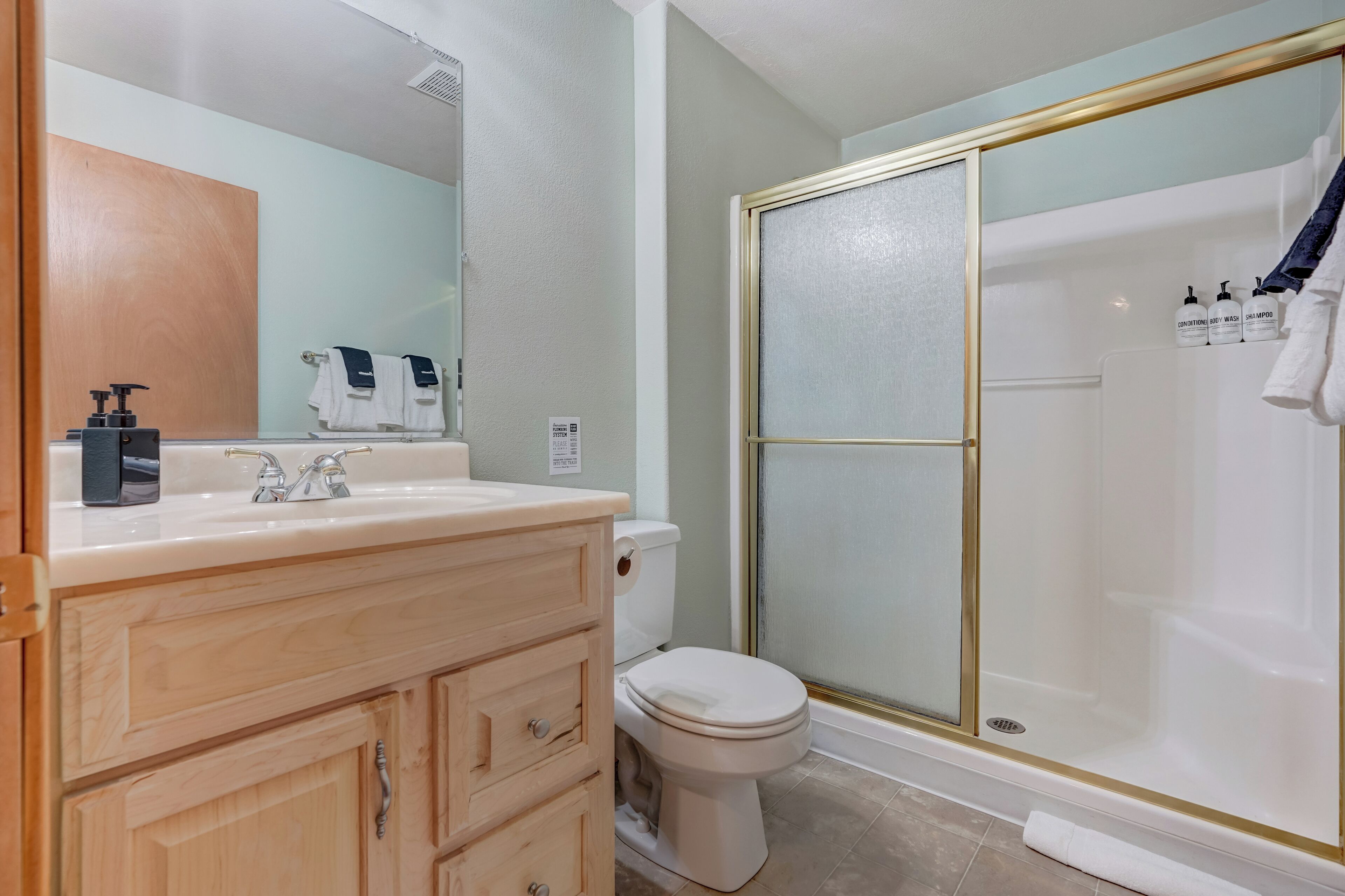 A bathroom with a wooden vanity, a toilet, a glass-enclosed shower, and toiletries on the sink.
