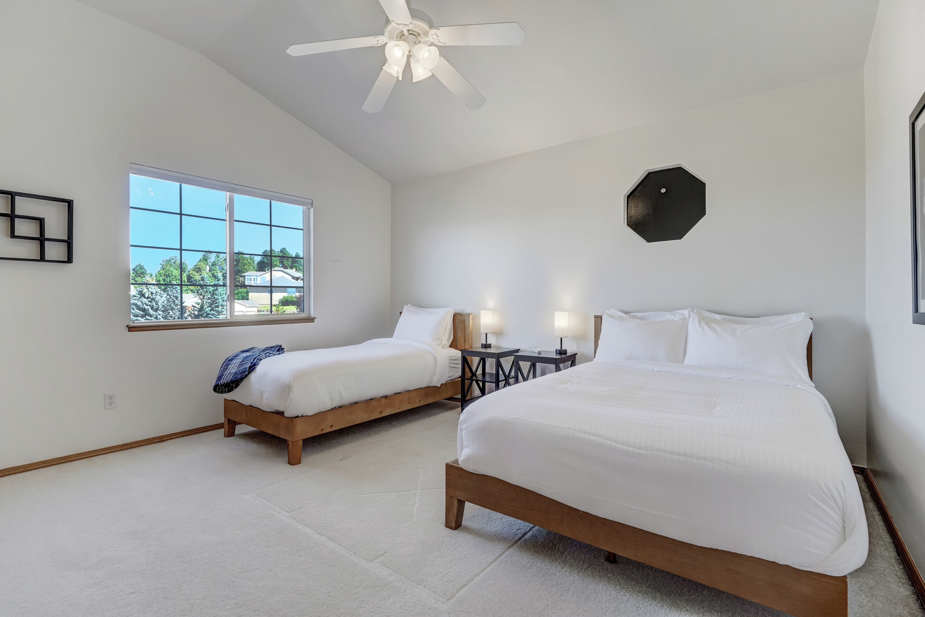 A bedroom with two beds, a ceiling fan, a window, and a small table with lamps.